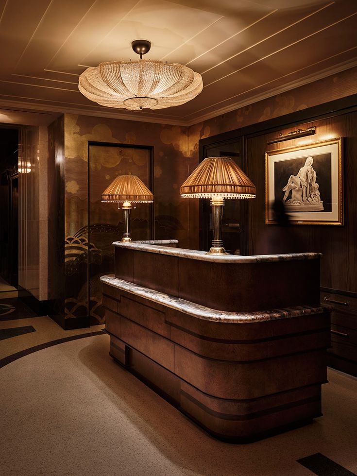 A softly lit hotel reception desk in burl wood and marble, with pleated table lamps and a deco ceiling light